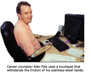 Career counselor Alan Fisk is seated and resting his prosthetic arms with hands made of stainless-steel hooks on his desk in front of him. He is smiling. His work station includes a computer with a touchpad designed to withstand the friction of his hands.