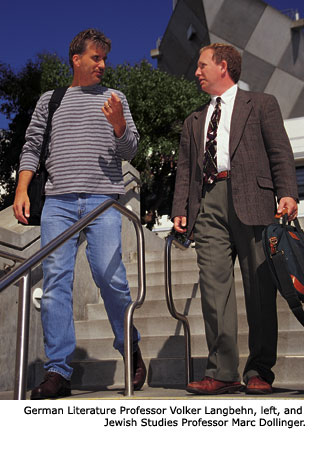 German Literature Professor Volker Langbehn, dressed in a long-sleeeved striped tee shirt and jeans, and Jewish Studies Professor Marc Dollinger, dressed in a suit and tie, are carrying on a conversation while walking down a set of stairs on campus outside the Cesar Chavez Studenr Center. Langbehn is using his hands to help him express a thought while Dollinger listens.
