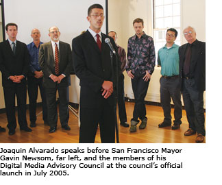 Joaquin Alavardo speaks into a microphone while Mayor Gavin Newsom and the members of his digital media advisory council stand in the background listening.