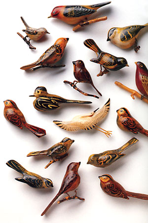 A photograph of 16 hand-carved wooden birds with tiny mesh legs. Each one is intricately detailed and looks different than the others.