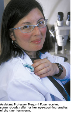 Assistant Professor Megumi Fuse in a white lab coat with a microscope behind her and a small green caterpillar attached to her lapel.