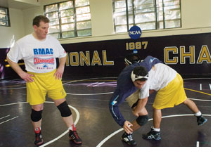 Wrestling coach Lars Jensen stands on the mats in a tee-shirt, shorts and knee pads and looks on as two student wrestlers try to pin one another down.