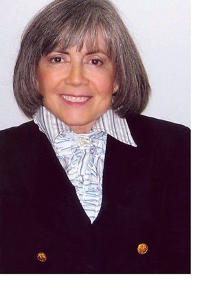 A smiling Anne Rice with shoulder length hair and a ruffled shirt buttoned at the neck and navy blazer with gold buttons.