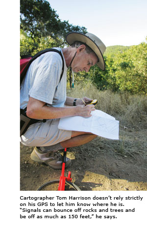 Cartographer Tom Harrison, with his walking stick, pen and compass handy, crouches at a trailside to make notations on a map.