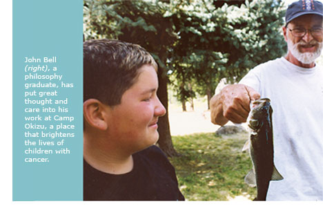 John Bell, wearing a baseball cap, holds a newly caught fish in his hand, earning an appreciative grin from a young camper.