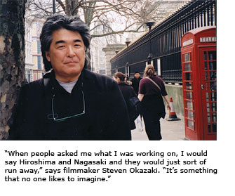 Filmmaker Steven Okazaki, glasses hanging from his neck, stands before a fence and British-style telephone booth.