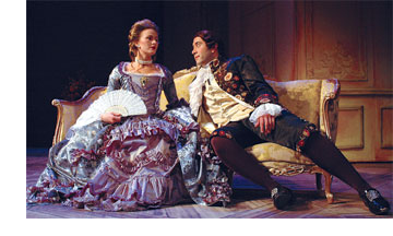 A woman sits stiffly on a couch, wearing a full-skirted gown of purple and lavender ruffles, a pearl choker and a white fan. A man seated near her leans in with interest. He wears a ruffled shirt, embroidered coat and wig of brown curls.