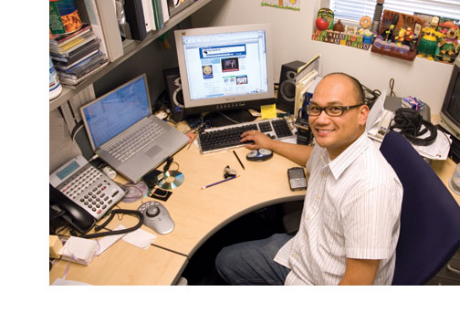 Two computers, a handheld device, a phone -- and fun desk toys -- surround a smiling Voltaire Villanueva as he fields admissions questions electronically.