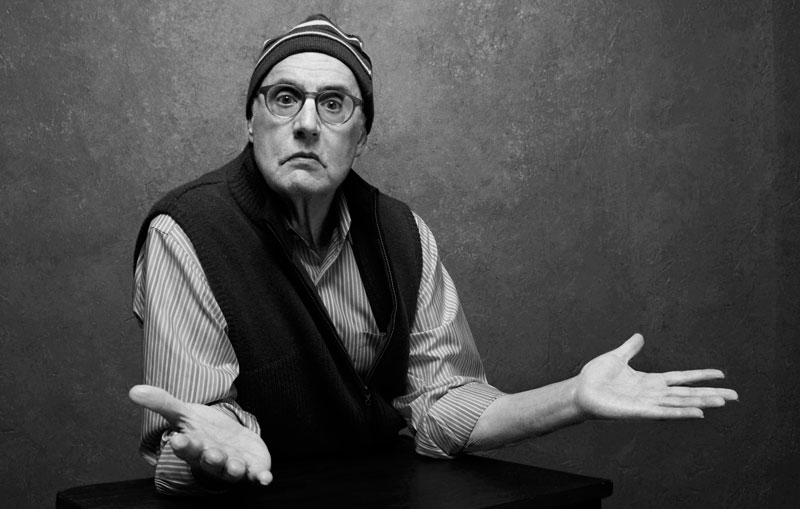 Jeffrey Tambor throwing his hands up in a questioning pose in front of a grey backdrop.