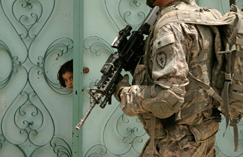 A girl looking through a gate in the middle east as a soldier walks by