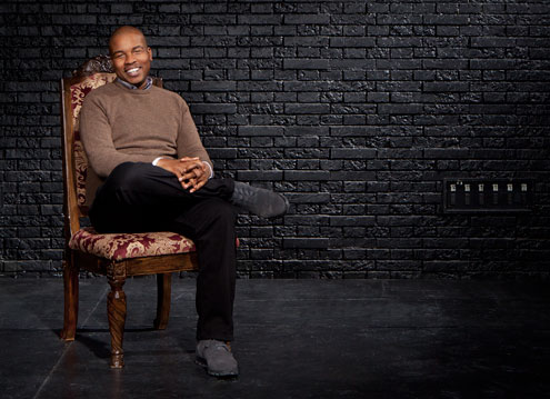 Marcus Gardley sitting on a wooden chair, smiling, with a black brick wall behind him.
