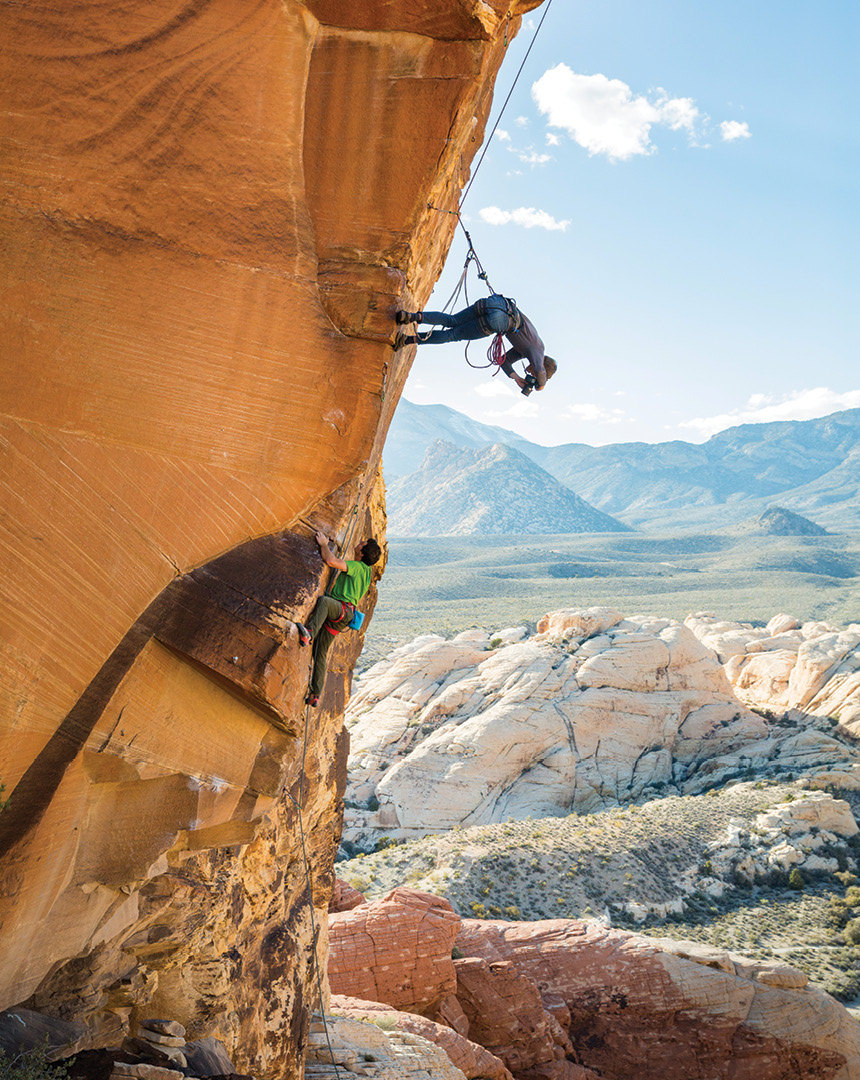 A photographer, strapped into a harness, taking a photo of a rock climber beneath him