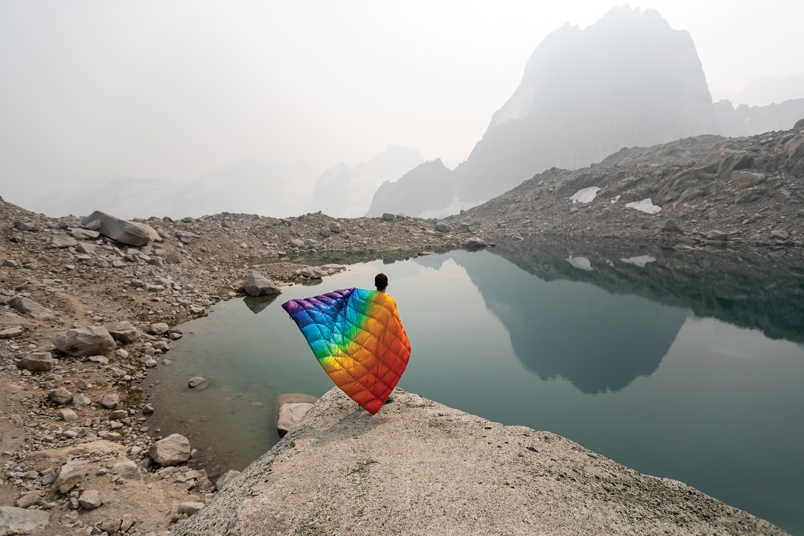 Man with a rainbow blanket, standing on a rock looking out into a barren lake
