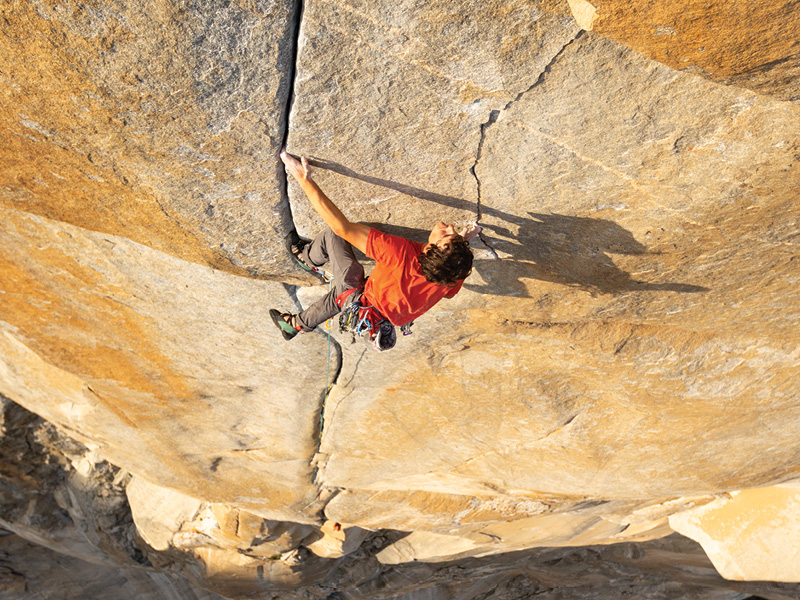 Guy in Orange shirt rock climbing up a very steep mountain face