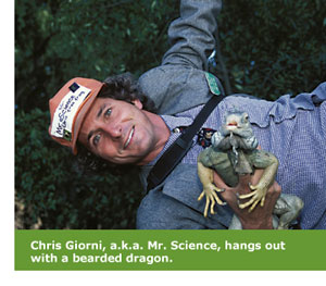 Alumnus Chris Giorni is stretched out on the ground and holding a bearded dragon in one hand. His cap says “Mr. Science.”