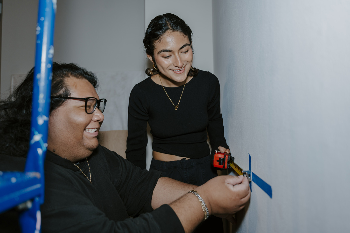 two students using a measure tape along a wall and marking it with painters blue tape